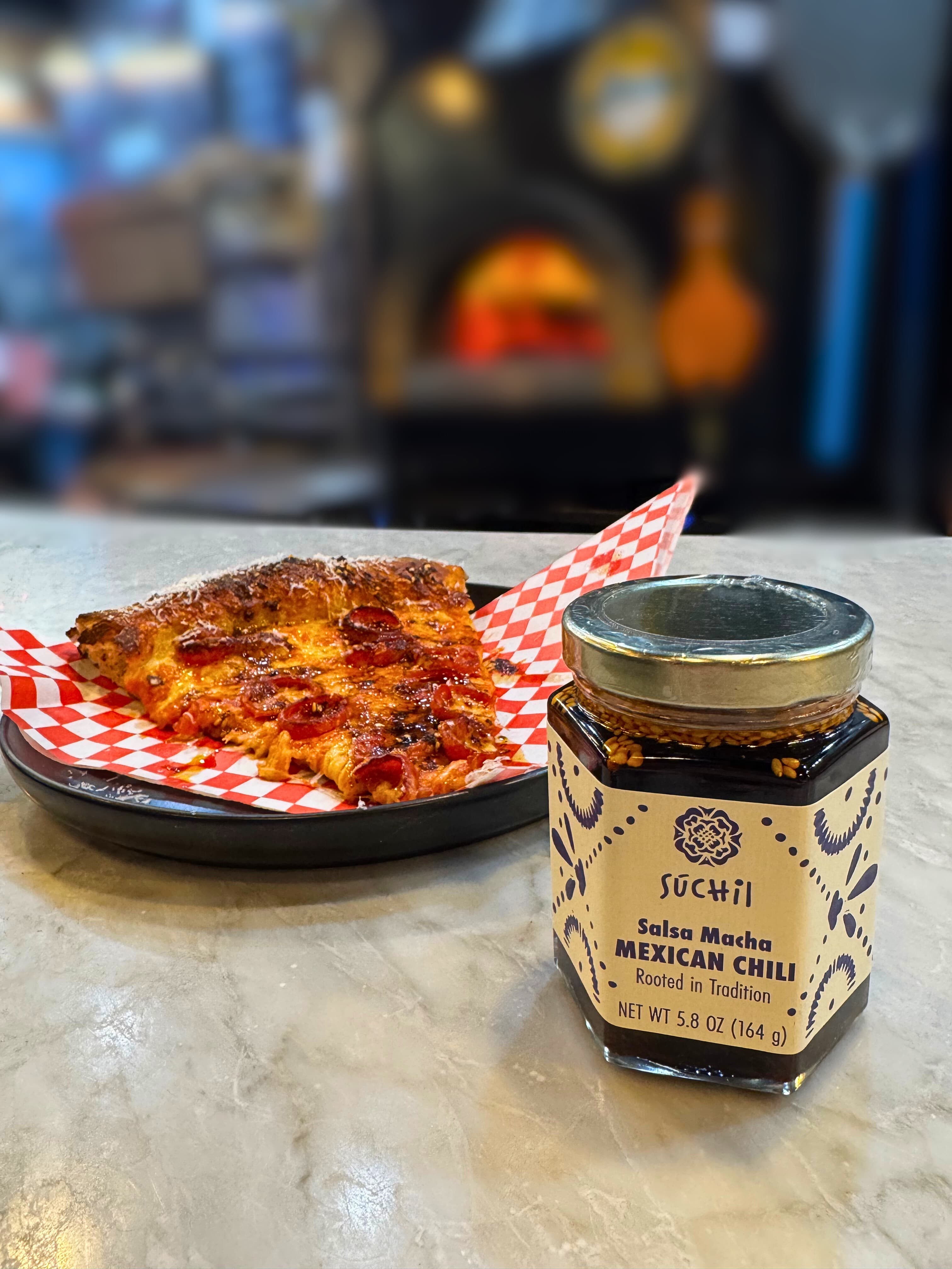 Súchil Made Salsa Macha jar beside a slice of pizza.